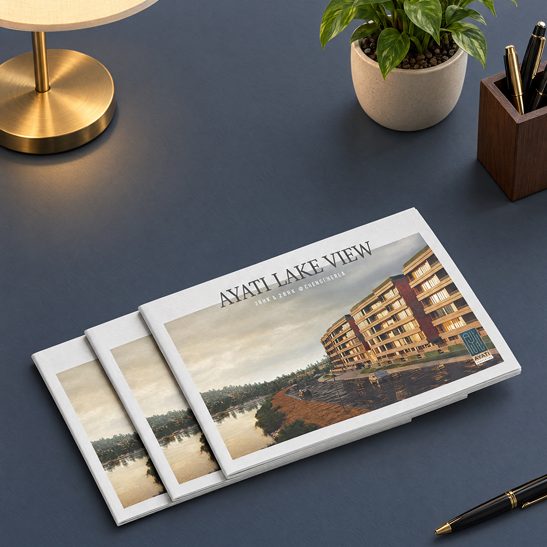 Ayati Lakeview: Brochure design mockup with three stacked brochures showing a lakeside apartment building, on a navy desk with a brass lamp, plant and pen
